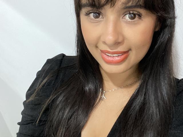 KeyklinOne has medium brown skin, dark straight hair, brown eyes, and is smiling with red lipstick. She's wearing a black top and a silver necklace with an "A" pendant.
