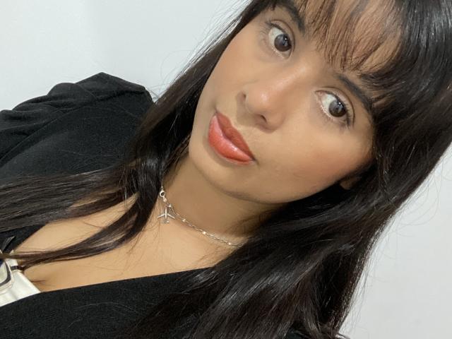 KeyklinOne with dark straight hair, bangs, brown eyes, and shiny red lipstick, wearing a black top and silver cross necklace, looking slightly to the side.