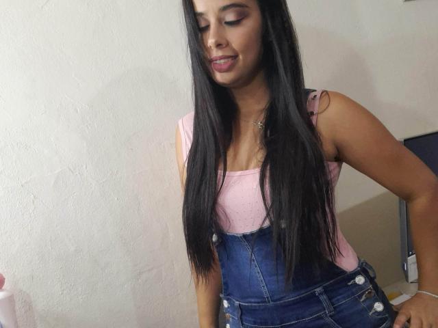 KeyklinOne with long black hair, pink tank top, and blue denim overalls, slightly smiling with eyes closed, standing in a room, showing a cute, sexy, and cute-ass look.