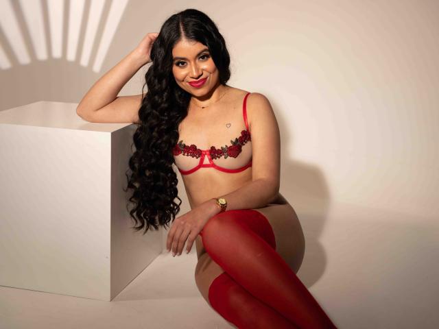 GabrielaRossa with long curly black hair, red lingerie with black lace, and red thigh-high stockings, sitting on a white cube, smiling, hand in hair, red lipstick, watch.