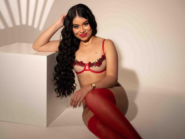 GabrielaRossa, con lunghi capelli neri ricci, indossa lingerie rossa con ricami floreali neri e collant rossi alti fino alla coscia, seduta su un cubo bianco, sorride, con una mano tra i capelli e un orologio al polso..