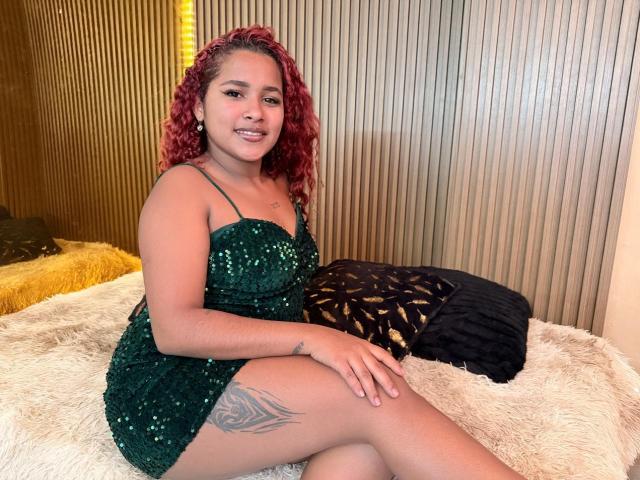 RubyLuna with red curly hair, green sequined dress, tattoo on thigh, sitting on white fur, smiling, dark and yellow pillows behind her. Her tits are small, pussy not visible.