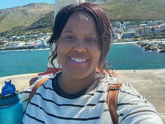 SweetBustyBee con la pelle scura, i capelli neri e un sorriso, che indossa una camicia a righe, spallacci marroni per uno zaino e una bottiglia d'acqua blu, in piedi vicino a una riva con montagne sullo sfondo..