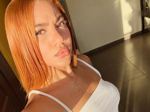 Close-up of KittyLara-hot with orange hair, brown eyes, and full lips. She wears a white spaghetti-strap top and a small cross necklace. Sunlight casts shadows on her face and chest. Her hair is straight and shoulder-length. The background is a dark, tiled floor with a yellow wall. Her expression is neutral, slightly pouting. The image has a warm, natural light.