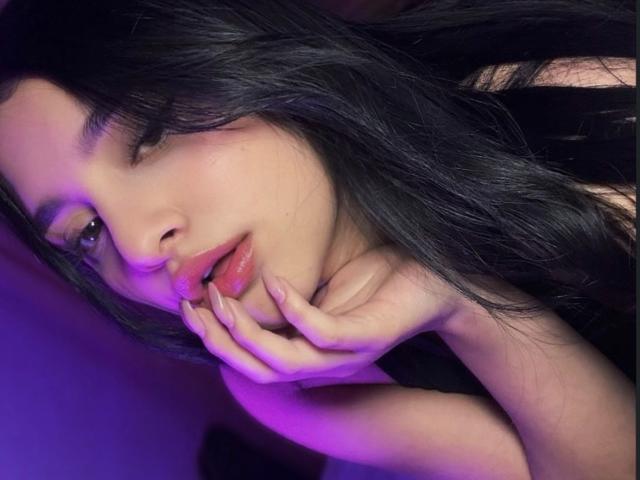MeganPaige with black hair, half-closed eyes, and purple-tinged skin, gently biting her finger, mouth slightly open, showing her pink lips and manicured nails.