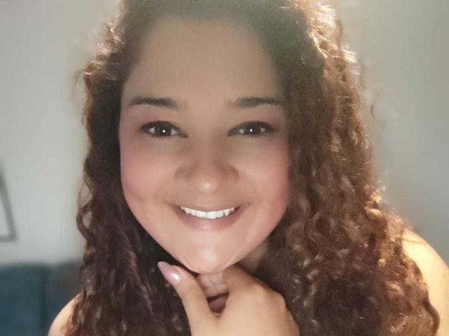 Close-up of NataliaWendys with curly brown hair, smiling at the camera. She has a light brown skin tone, dark eyebrows, and brown eyes. Her lips are slightly parted, showing white teeth. She is touching her chin with her right hand. The background is blurred, focusing on her face. Her hair is thick and textured, and she has a subtle blush on her cheeks. The image has a soft, natural light, highlighting her facial features. Her expression is relaxed and friendly.