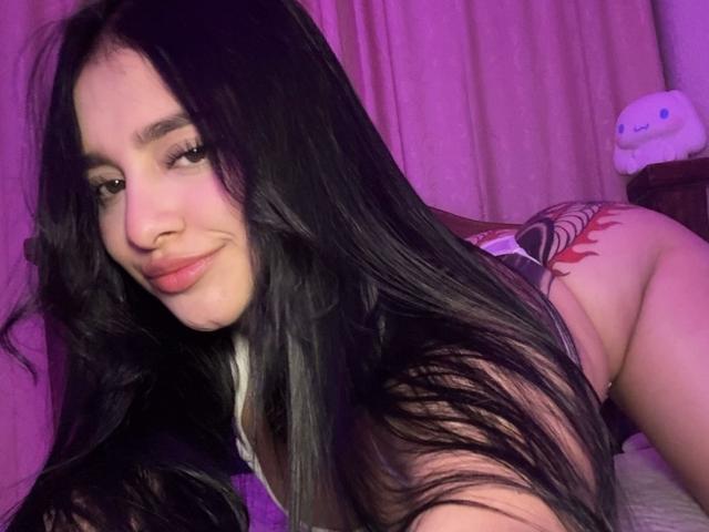 MeganPaige with long black hair, smiling, showing a red tattoo on her right ass cheek, lying on a bed, purple lighting, looking at the camera.