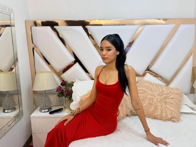 SweetPalmer with long black hair in a red dress, sitting on a white bed with a fluffy pink pillow, her ass and tits slightly visible, looking at the camera.
