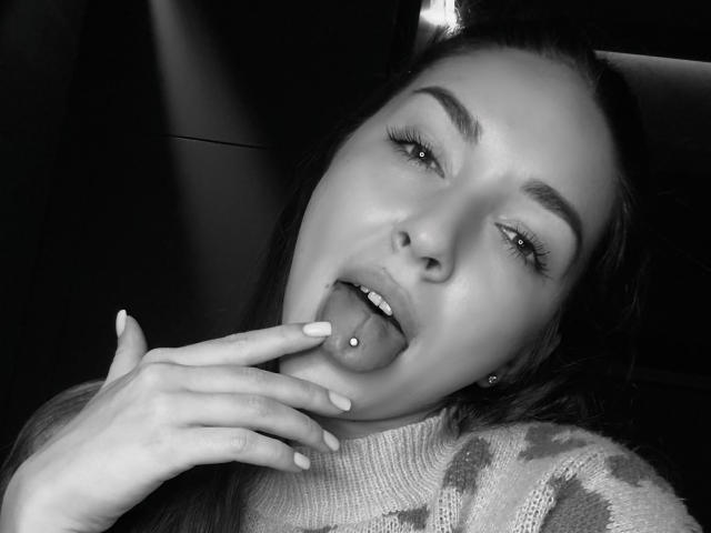 AquaLady's face in a black-and-white photo, tongue out, showing a tongue piercing, with fingers touching her tongue. She's wearing a patterned sweater, looking sexy and cute.