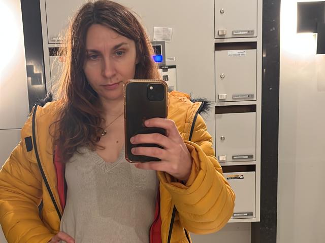 JasmineGlamour stands in a mirror selfie, holding a black smartphone with a gold edge. She has wavy, shoulder-length brown hair and a neutral expression. She wears a yellow puffer jacket with blue fur trim and a beige, slightly wet, V-neck shirt. Her left hand rests on her hip. The background shows a white and black wall with shelves and equipment. The lighting is bright and even. The composition focuses on JasmineGlamour's reflection and her upper body.