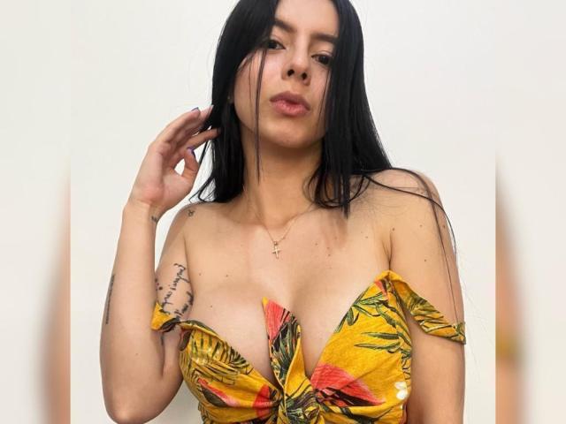IsabellaJames with long black hair, pouty lips, yellow off-shoulder top with red and green pattern, small cross necklace, tattoo on left arm, hand in hair, light skin.