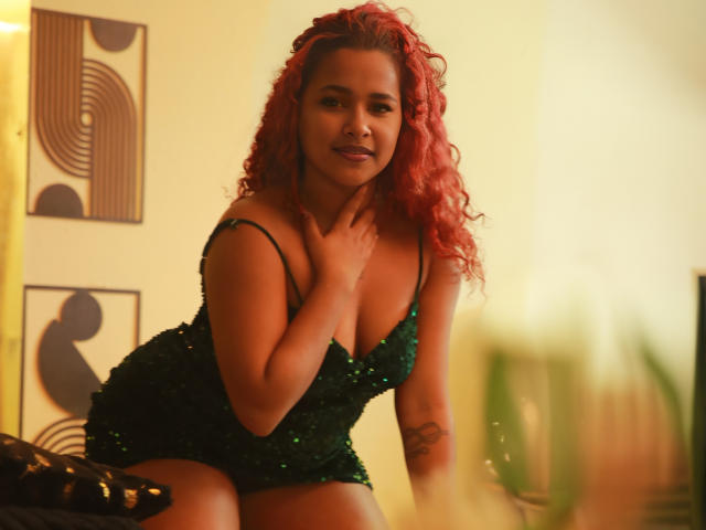 RubyLuna with red curly hair, wearing a green sequined dress, touching her neck, showing her tits and a tattoo on her left arm, in a warmly lit room.