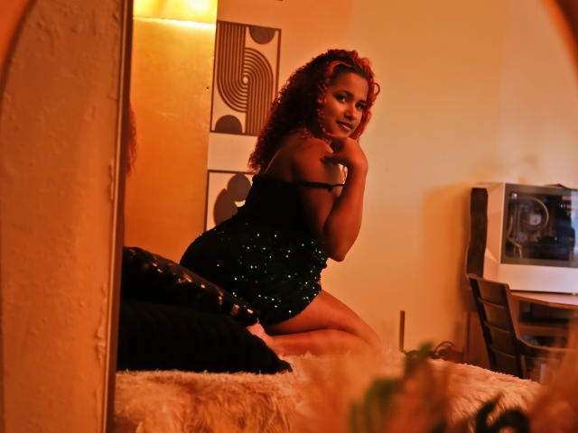 RubyLuna with curly red hair, wearing a black, sparkly, short dress, kneeling on a fluffy bed, hand on chin, looking seductively at the camera, warm orange lighting.