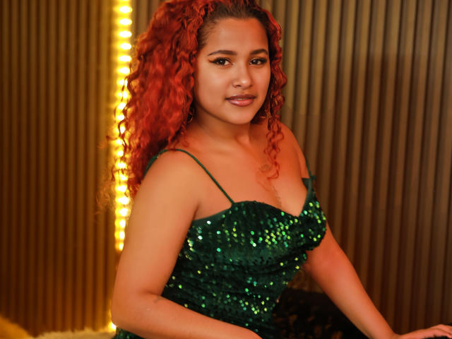 RubyLuna with red curly hair, green sequined dress, and cute smile, showing her boobs and ass, sitting in warm, yellow-lit room. Her brown eyes are attractive and confident.