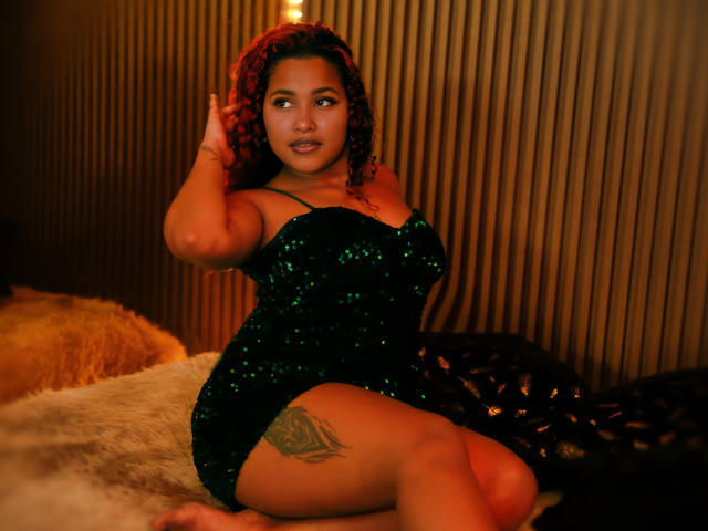 RubyLuna with curly red hair, green sequined dress, and tattoo on her thigh, sitting on a furry bed, hand in hair, looking to the side, sexy and confident.