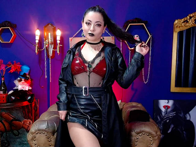 LunaTikka with black lipstick, red bra, and black leather dress, holding her long black hair, showing her tits through sheer fabric, standing in front of a blue wall.
