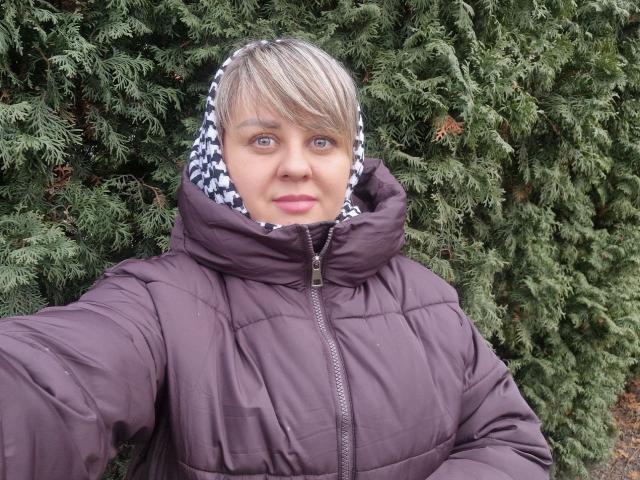 VeronicaSvitt with short blonde hair, blue eyes, wearing a puffy purple jacket with a black-and-white checkered hood, standing in front of green pine trees. Boobs are not visible.