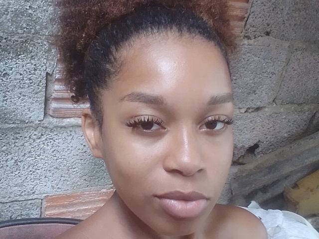 PoneiZinha has brown curly hair, brown eyes with long eyelashes, and brown skin. Her small, round, shiny ass is partially visible in the lower right corner.