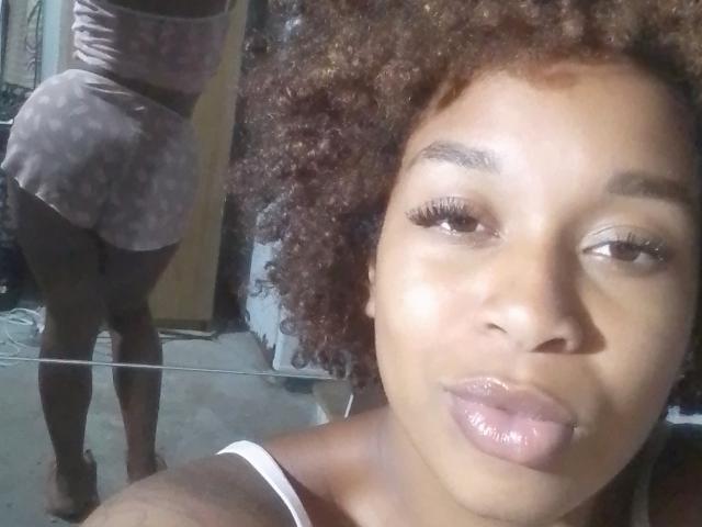 PoneiZinha with curly brown hair, glossy pink lips, wearing a pink floral skirt, showing her back and big black ass in a mirror selfie. PoneiZinha's face is in the foreground.
