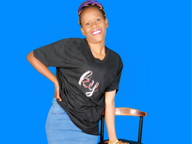 Sharice met donkere huid, roze lippenstift, een zwart shirt, de tekst "By", blauwe jeans, leunend op een stoel, een paarse hoofdband, lachend, met handen op de heupen, op een helderblauwe achtergrond..