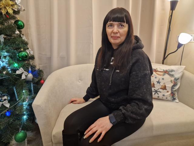 BlairFlepp, middle-aged woman with dark brown hair, sitting on beige couch, wearing black hoodie and black pants, next to decorated Christmas tree with green and blue ornaments.