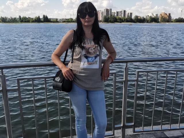 BlairFlepp with long black hair, wearing sunglasses, a gray graphic tee, blue jeans, and a black shoulder bag, standing by a metal railing by a rippling water body.