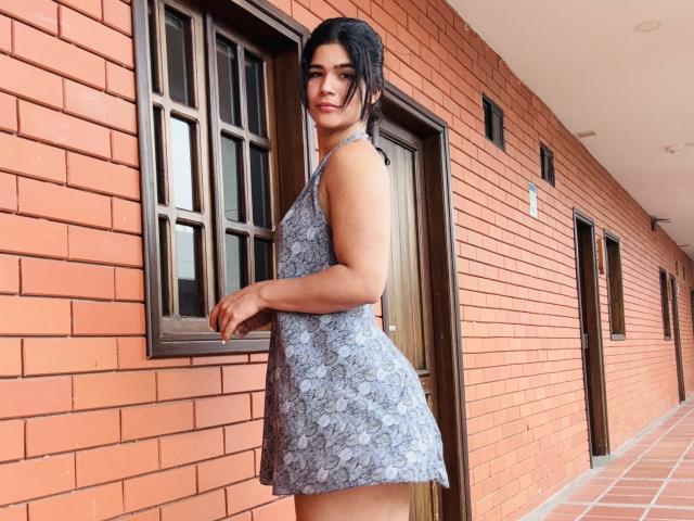 ScarlletZhen with dark hair, wearing a grey floral dress, standing against a red brick wall, showing her ass and looking back with a slight smile.