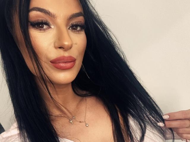 KinkyAmelie has long black hair, brown eyes, and full red lips with a small beauty mark on her right cheek. She's wearing a necklace and a pink top.