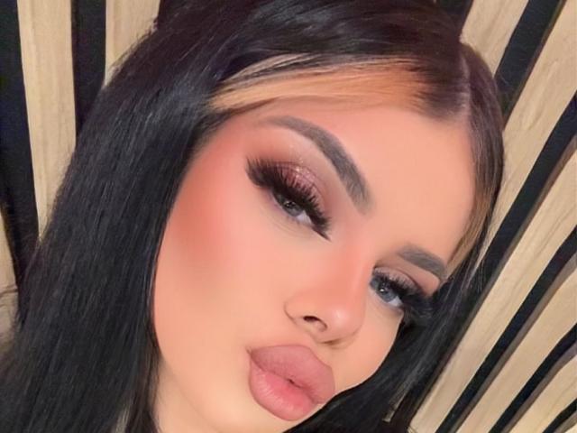 IvyRaven's face with puffy pink lips, dark long hair, heavy black eyelashes, and pinkish-red makeup, looking slightly to the side with a subtle smile.