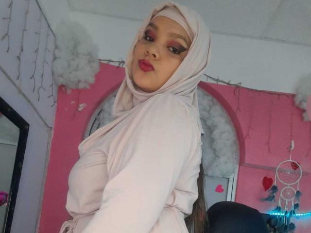 AzaharaMahmud with pink hijab, pink long-sleeve top, looking back, showing her tits, red lipstick, and dark eyeliner, in a pink room with white fluffy clouds.