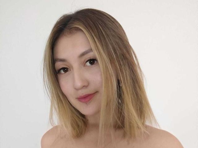 AngelaTorres has blonde, shoulder-length hair, light skin, brown eyes, and a subtle smile with pink lips. She's topless, showing her bare shoulders and upper chest.