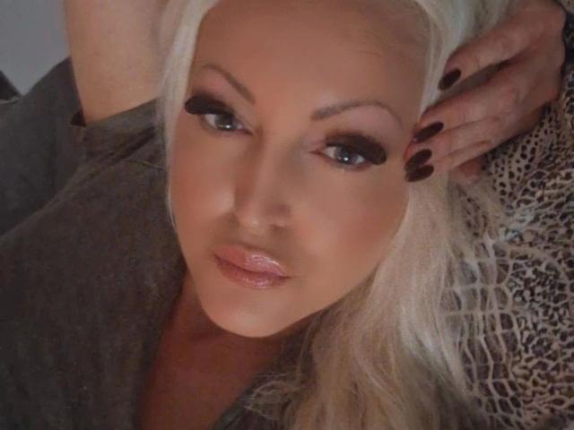 KimLayne-hot with platinum blonde hair, dark eyeliner, and glossy pink lips, has her hand on her head, showing dark nails. She's wearing a grey top with a leopard print.