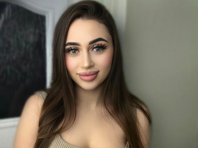 OliviAngel has long brown hair, green eyes, full pink lips, and is wearing a beige top. Her tits are partially visible. She's smiling softly with well-defined eyebrows.