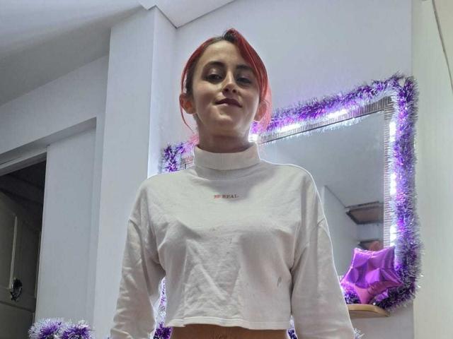 IvanaNovoa with red hair, wearing a white long-sleeve shirt, showing small tits, standing in a room with purple tinsel, smiling at the camera.