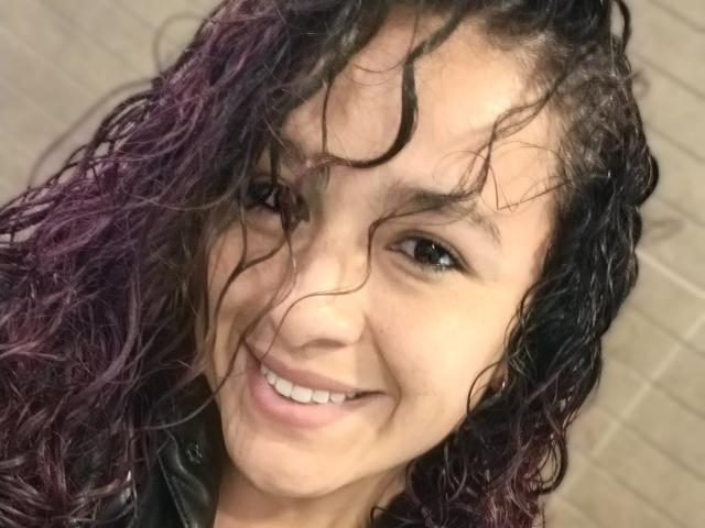 LilibethSexy has curly, wet black and purple hair, a warm smile, and light brown skin. She's wearing a black leather jacket, looking cute and sexy.