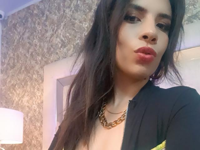 VioletaStone, met lang donker haar, volle rode lippen, gekleed in een zwart top met rits en een gouden ketting, kijkt in de camera met lichtelijk gefronste wenkbrauwen..