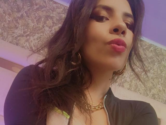 VioletaStone with wavy brown hair, hoop earrings, gold chain, black top, and pink lipstick, pouting her glossy red lips. Her medium tits are slightly visible.