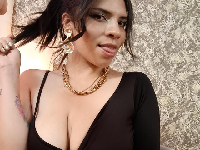 VioletaStone with big tits, wearing a black low-cut top, gold chain necklace, and large crescent moon earrings, holding her hair, looking at the camera with a slight smile.