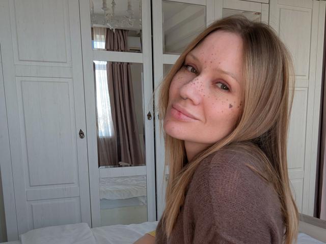 RamonaLust-hot tiene pecas, cabello rubio largo y una cara bonita, lleva una camisa marrón, con una ligera sonrisa, en un dormitorio con puertas de armario blancas..