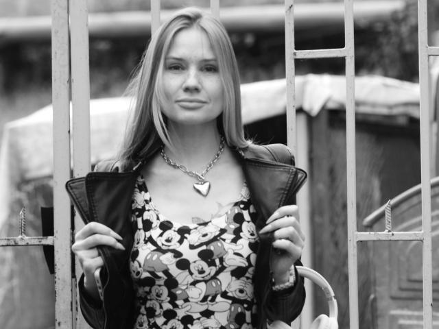 RamonaLust-hot está de pie frente a una puerta de metal, vestida con un vestido floral y una chaqueta de cuero, sosteniendo los bordes de la chaqueta, con un collar con forma de corazón y una ligera sonrisa..