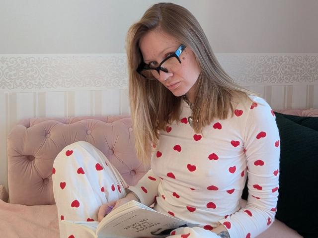 RamonaLust-sensual, con gafas, cabello rubio, pijamas blancos con estampados de corazones rojos, leyendo en un sofá rosa con adornos, piernas cruzadas, almohada verde a la derecha, sonriendo ligeramente, relajada..