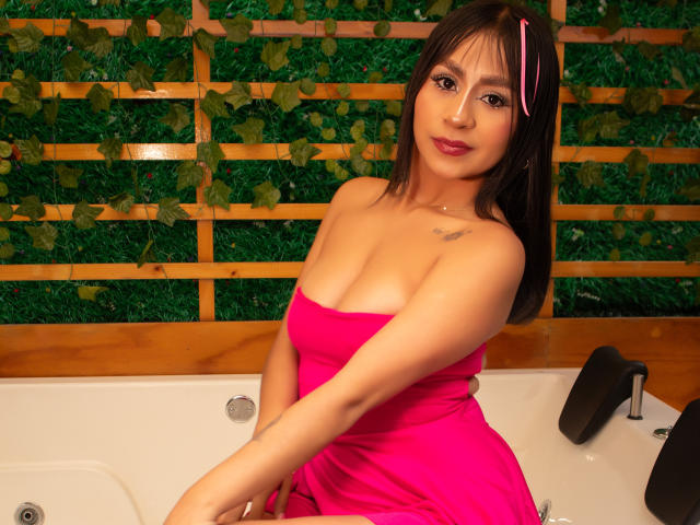 KimiNaru mit mittlerer Brustgröße, in einem pinken Kleid und mit Haarklammer, sitzt in einer Badewanne. Im Hintergrund sind eine grüne, blattreiche Wand, Holzlatten und schwarze Kopfstützen zu sehen..