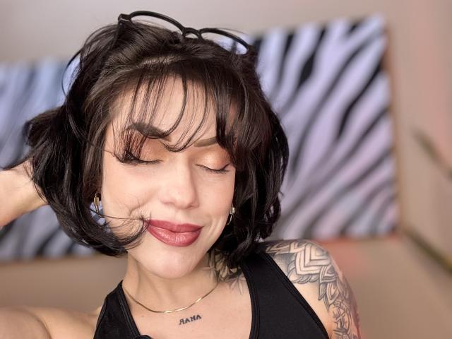 HannaBorbon with short black hair, closed eyes, and red lipstick, wearing a black tank top, showing a tattoo on her right shoulder, and a necklace. Her ass is not visible.