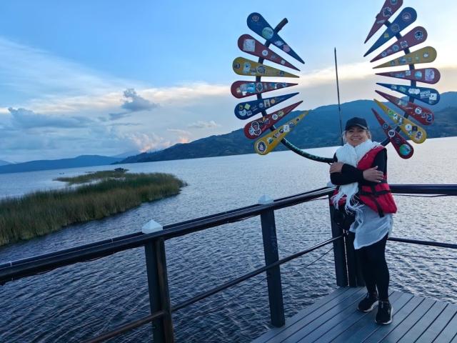 LadyLucky stands on a wooden dock, holding a fishing rod with a colorful, windmill-shaped lure. She wears a black cap, white shirt, black jacket, and red backpack. Her black pants and black shoes contrast with the dock's grey planks. The background features a calm lake, grassy marsh, and distant, blue-hued mountains. The sky is partly cloudy with a soft, early evening light. The lure's bright colors—yellow, blue, red—stand out against the natural landscape. LadyLucky's relaxed, smiling expression conveys a sense of calm and enjoyment.