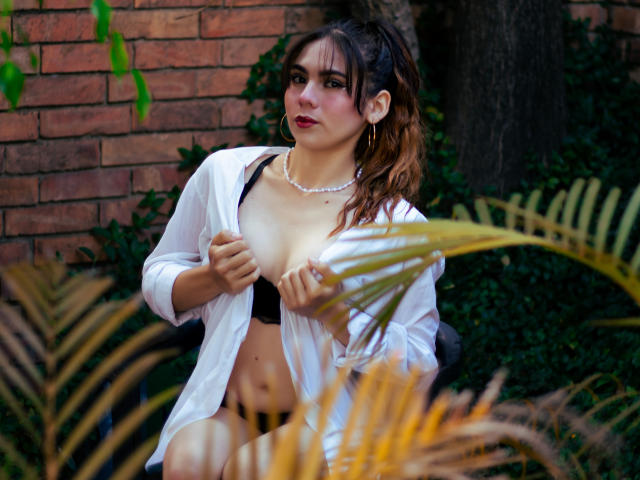 LindaPerez with brown hair in a ponytail, wearing a white shirt open to reveal black bra, exposing her tits, sitting against a brick wall, green necklace, looking off-camera.