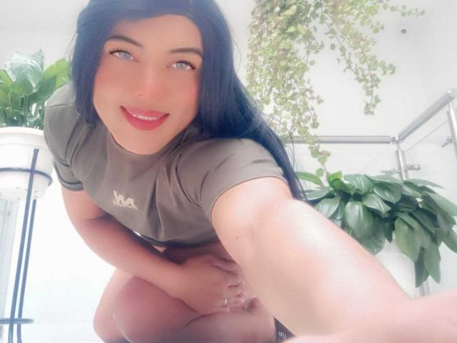 CharlotteSweetX, crouching with long black hair, red lipstick, and green eyes, wearing a grey shirt, smiling at the camera, showing her ass in a bright room with plants.