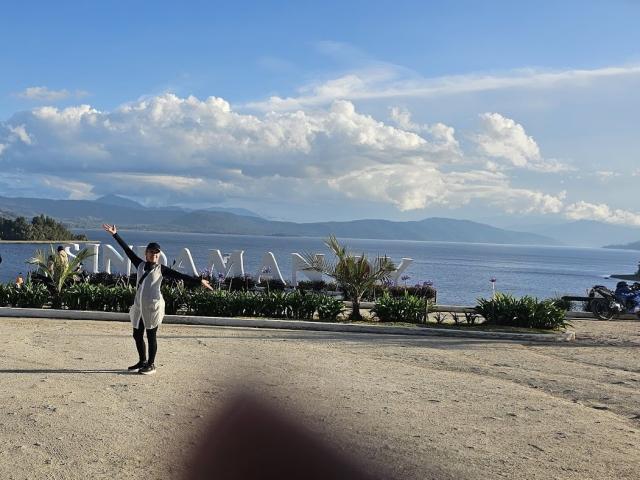 LadyLucky, standing on a gravel path, arms raised, wearing a black and white dress, facing a waterfront with large white letters and palm plants, under a blue sky.