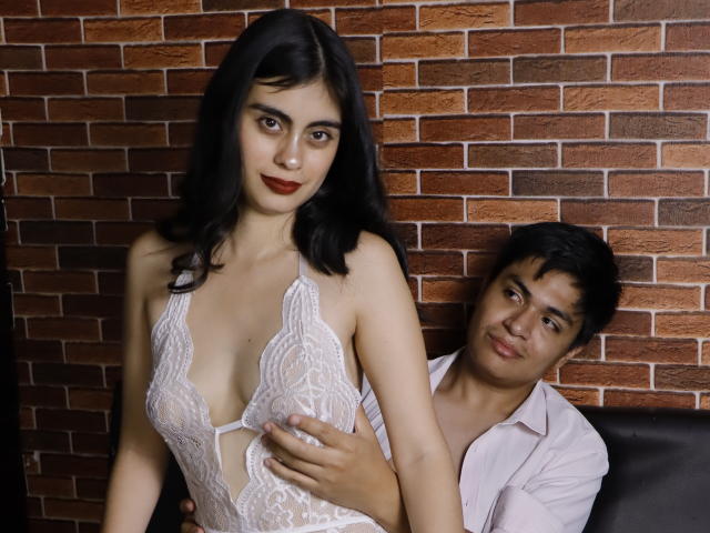 SofiNdLogan with dark hair, wearing a white lace lingerie dress, has small tits, and a guy in a pink shirt, with a hand on her tits, looking at her.