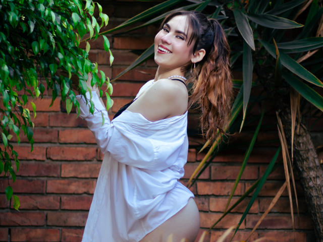 Linda Perez, avec des cheveux bruns ondulés attachés en queue de cheval, souriante, portant une chemise blanche à une épaule, laissant apparaître son soutien-gorge noir et ses fesses nues, se tient près de feuilles vertes et d'un mur de briques..