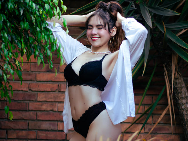 LindaPerez with her hands in her brown hair, wearing a white open shirt, black lace bra, and matching panties, smiling with red lipstick, showing her tits and pussy.